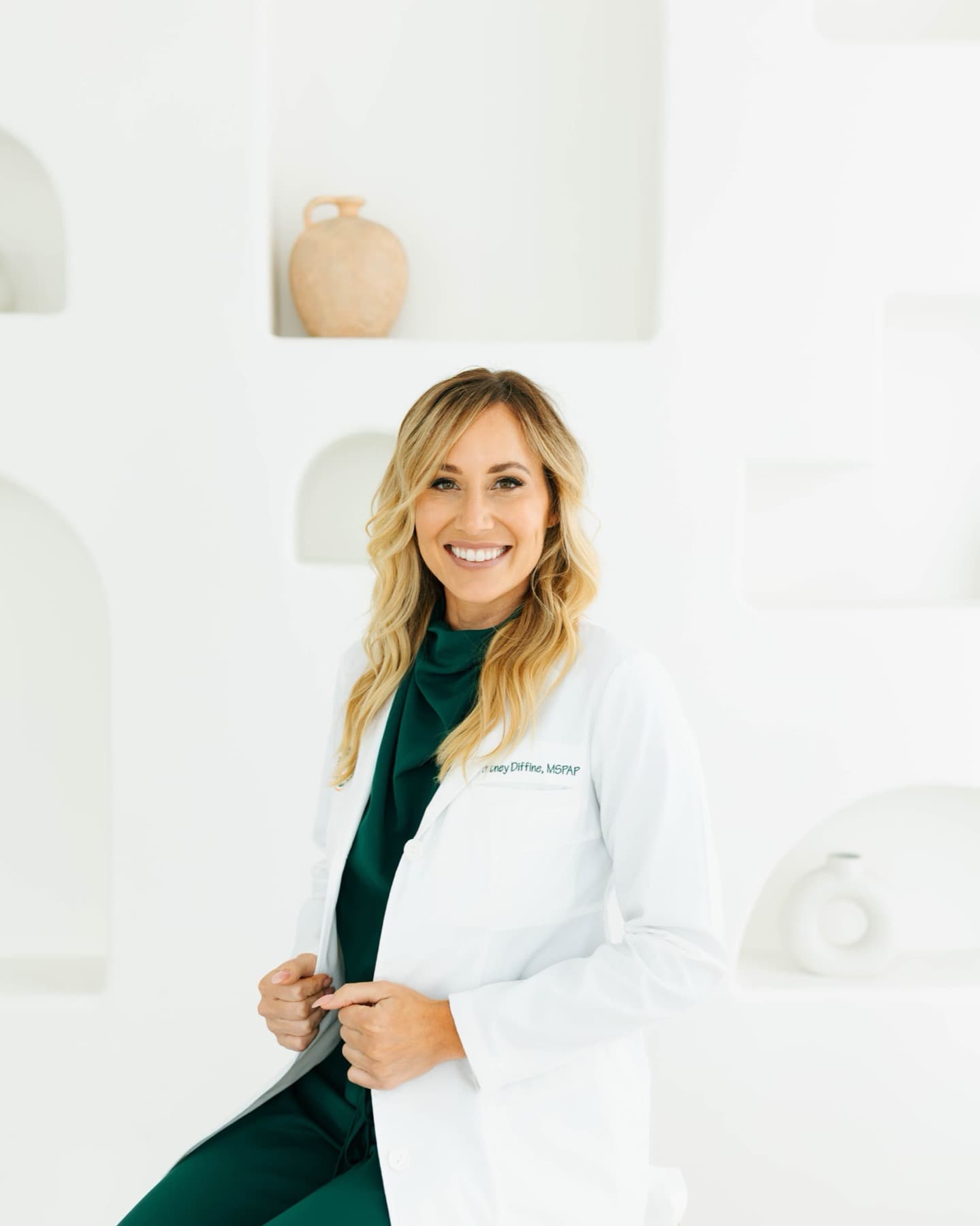 Brittney Diffiné, PA-C, in a white lab coat over emerald scrubs, smiling to camera